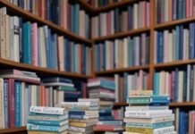 The Best Kindle Models Compared – Which One Is Right for You? Tabby cat swats bookmark on rickety table with Kindles, tiny bookshelves, pastel hues.